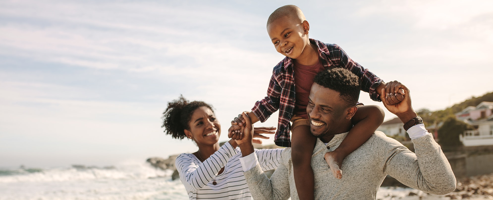 A smiling mom and dad with his son on his shoulders walk on a beach during vacation.