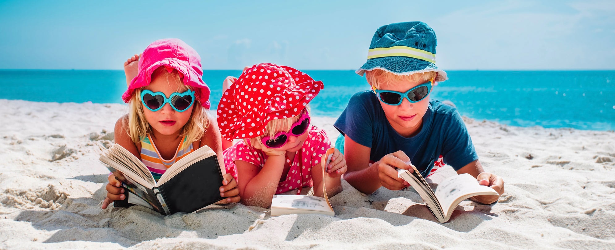 Three kids laying on the beach reading books.