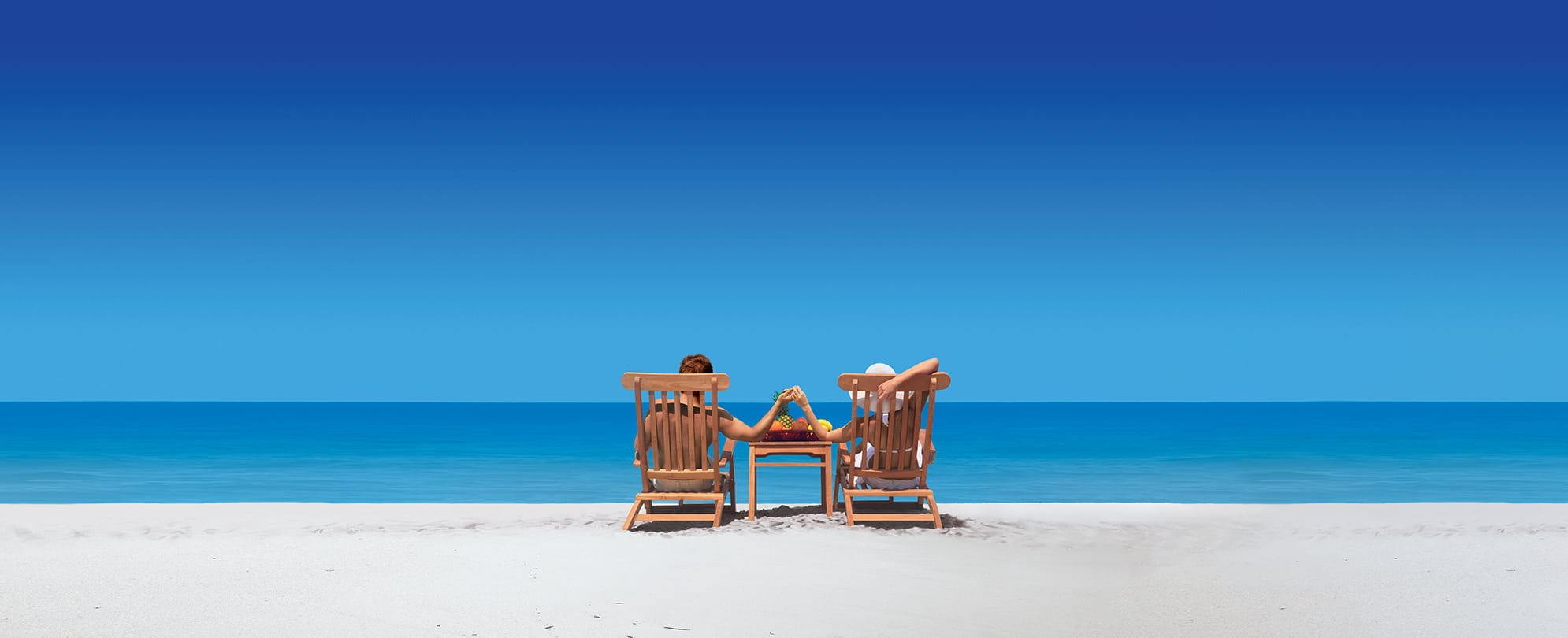 Couple on the beach facing the ocean sitting in beach lounge chairs holding hands over a side table 