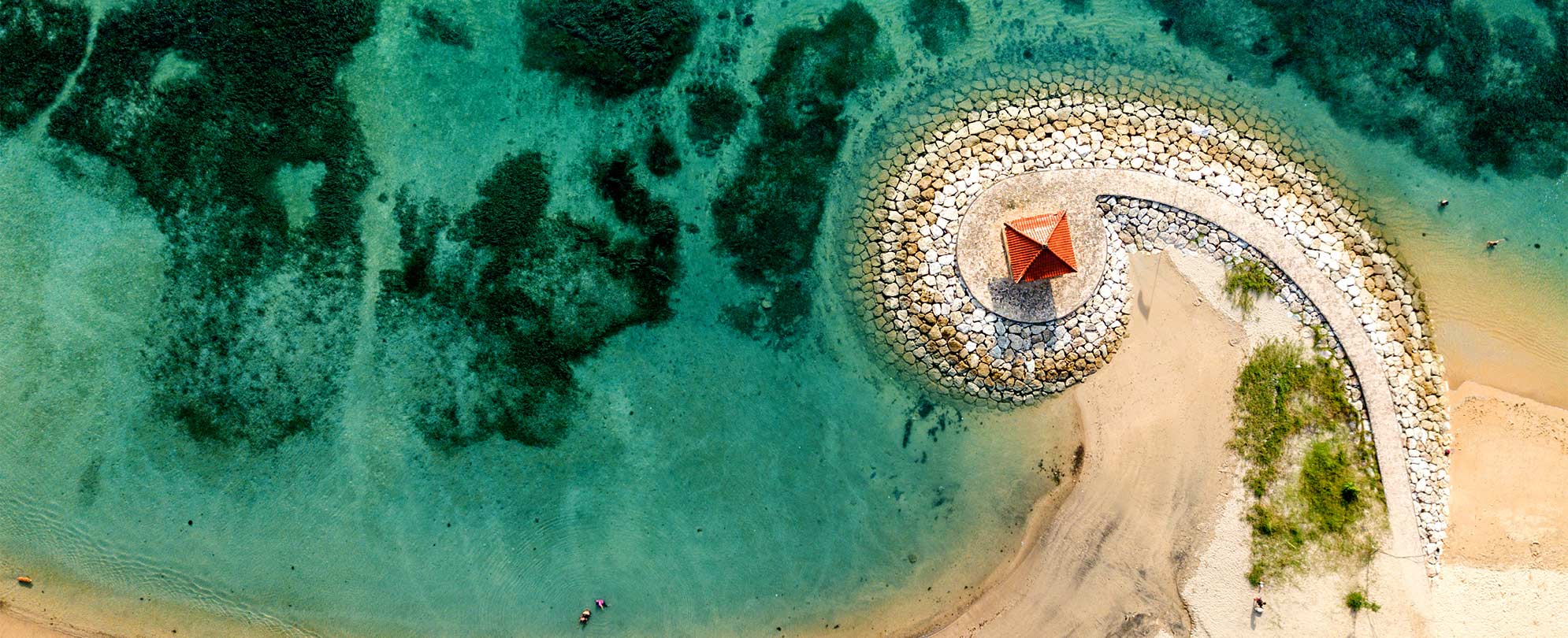 A bird's-eye view of a spiraling stone walkway on a beach by a crystal blue ocean.