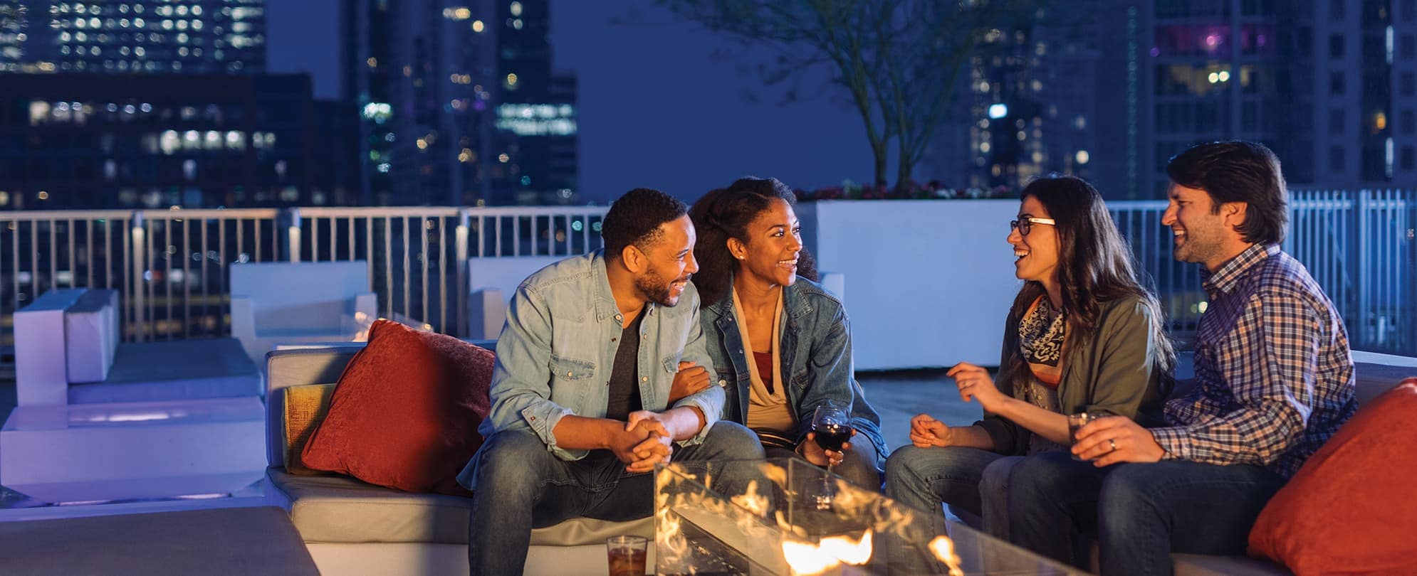 Two couples having a conversation at an outdoor rooftop lounge in front of a fire pit 