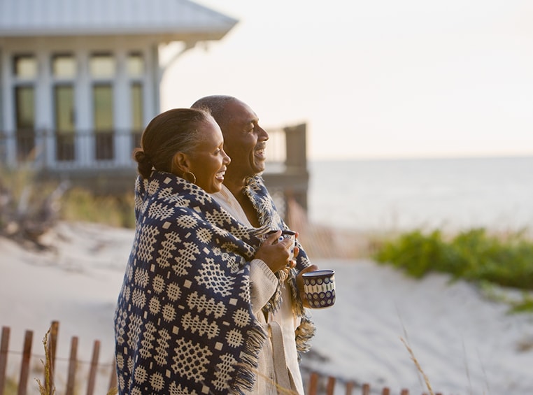 An older couple wrapped up in a blanket smile, looking out over the ocean.