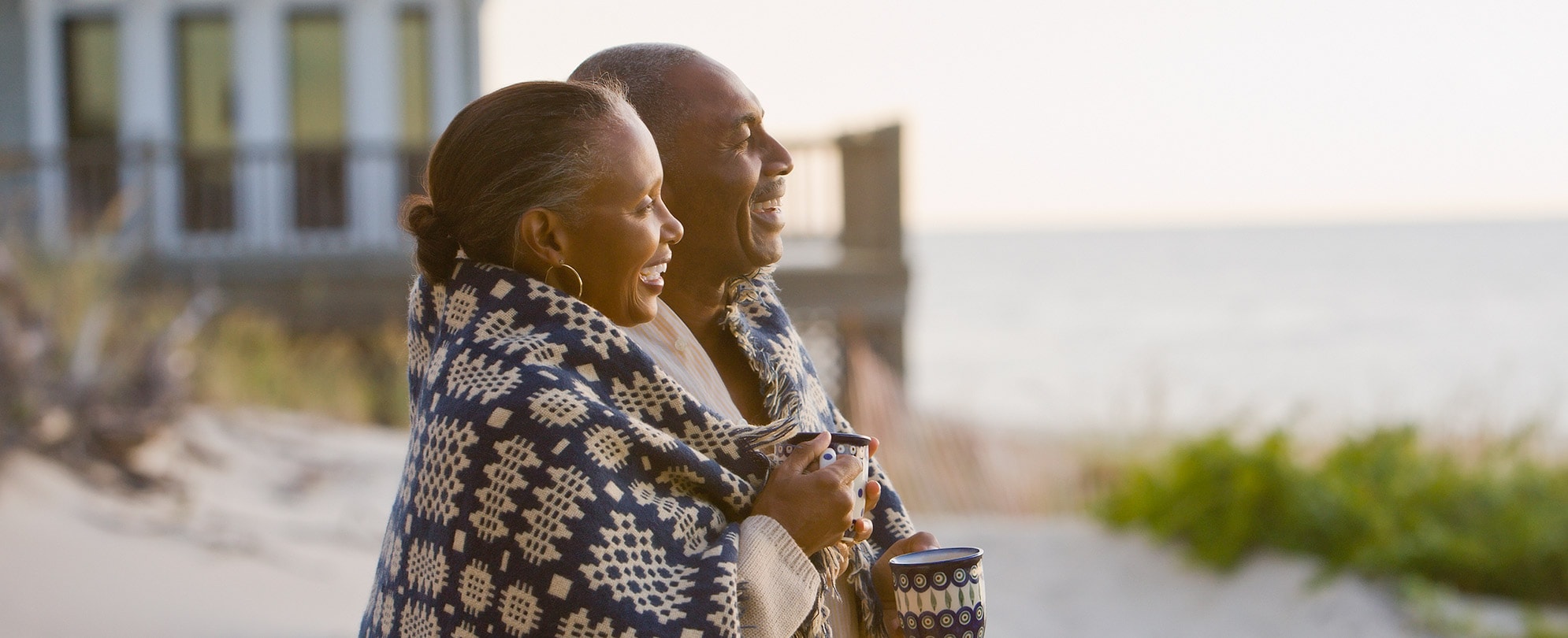 An older couple wrapped up in a blanket smile, looking out over the ocean.