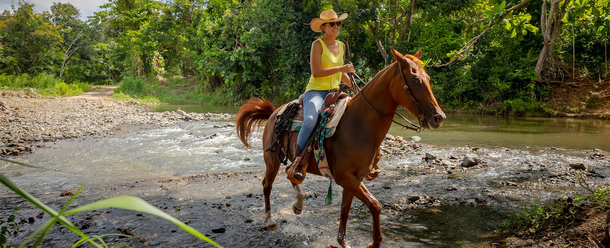 Woman wearing a yellow tank top and a wide-brimmed hat riding a horse through a rocky river. 
